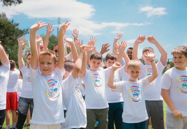 A group of cheerful children in white T-shirts raising hands in a park during summer.