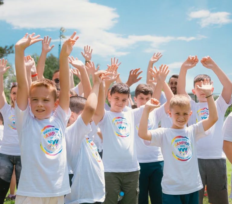 A group of cheerful children in white T-shirts raising hands in a park during summer.